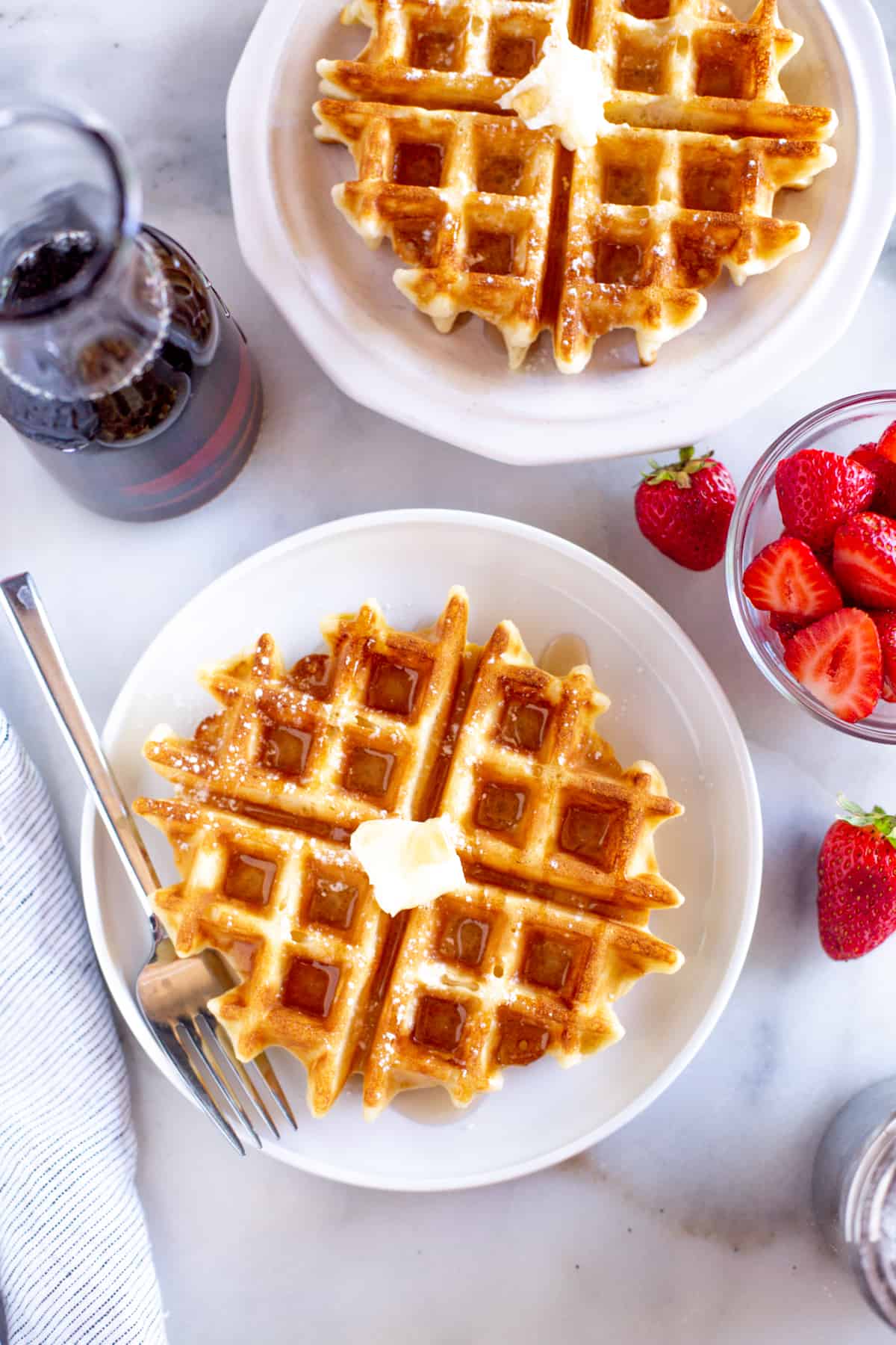 Belgian waffles on plates with butter, syrup, and powdered sugar next to a bowl of strawberries.