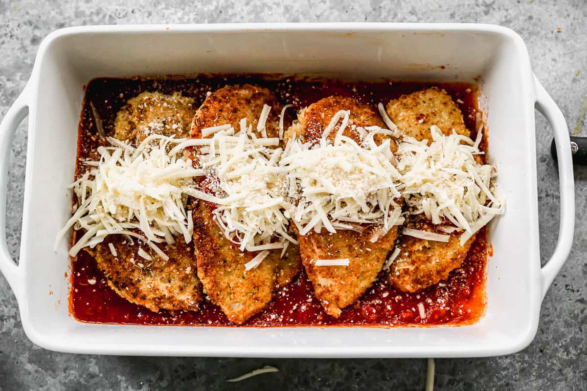 Four breaded chicken breasts in a white baking dish with marinara sauce and mozzarella cheese for Chicken Parmesan.