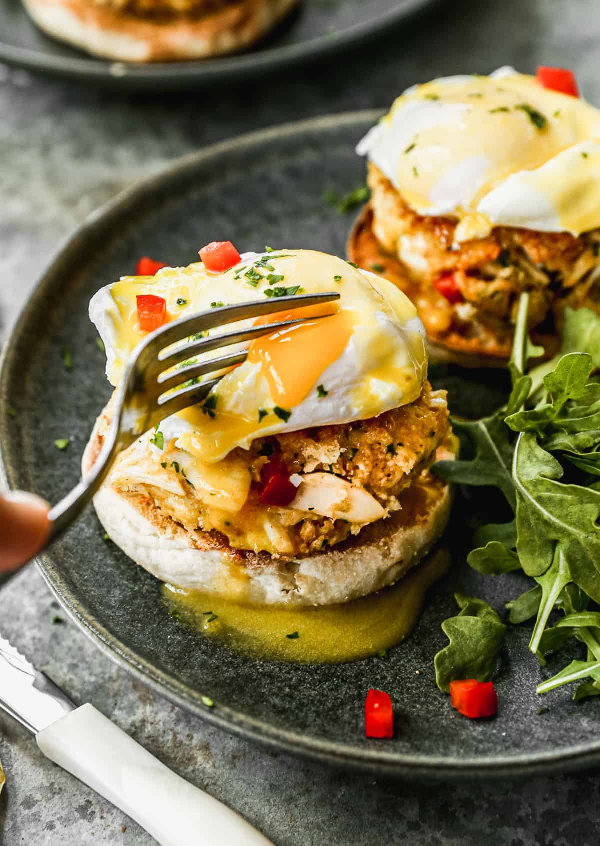 Crab Cake Benedict being cut into with a fork on a plate next to some greens.
