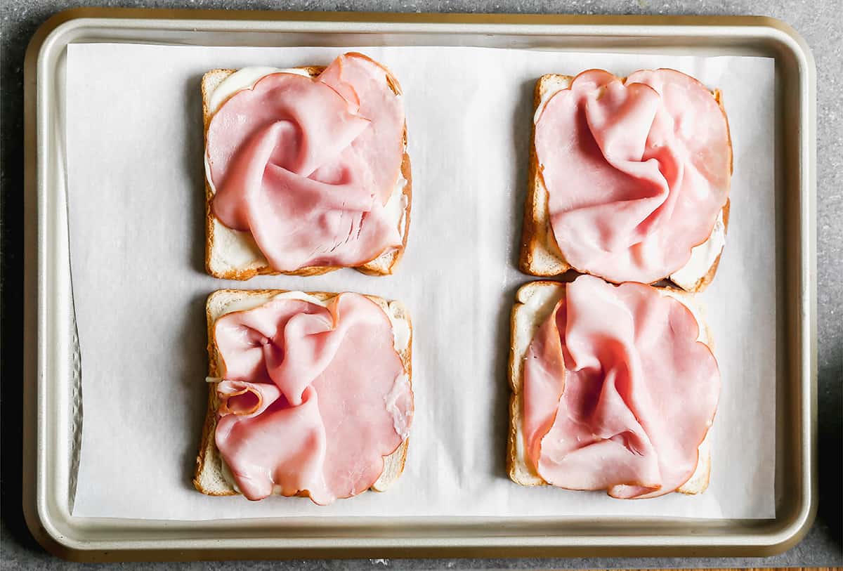 Four slices of bread on a baking sheet with béchamel sauce and a piece of ham on top.