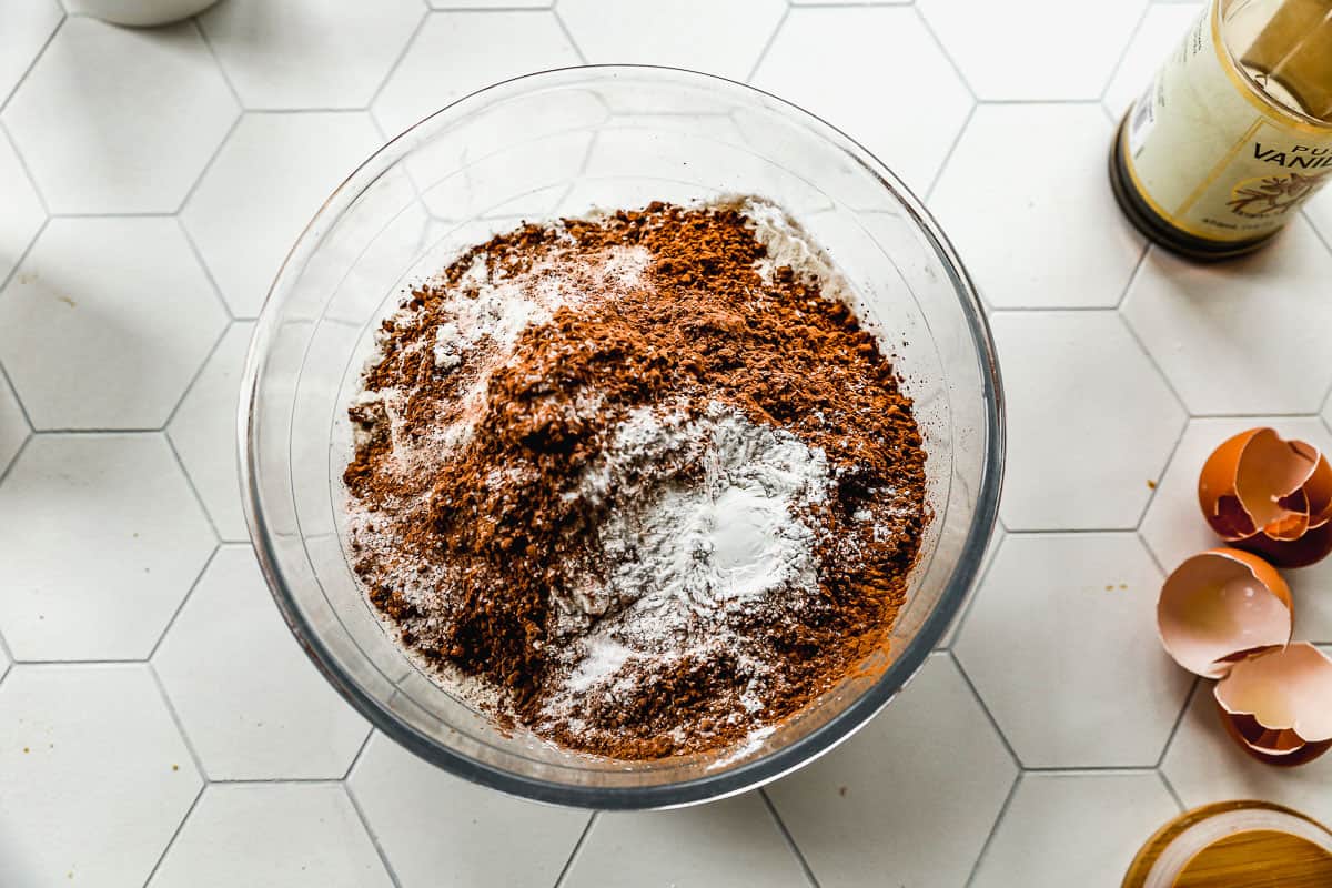 A glass mixing bowl with sugar, flour, cocoa, baking powder, baking soda, and salt for a homemade chocolate cake.
