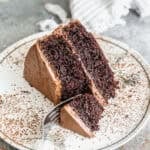 A slice of the best chocolate cake recipe served on a white plate with a sprinkle of cocoa powder and a fork cutting into it.