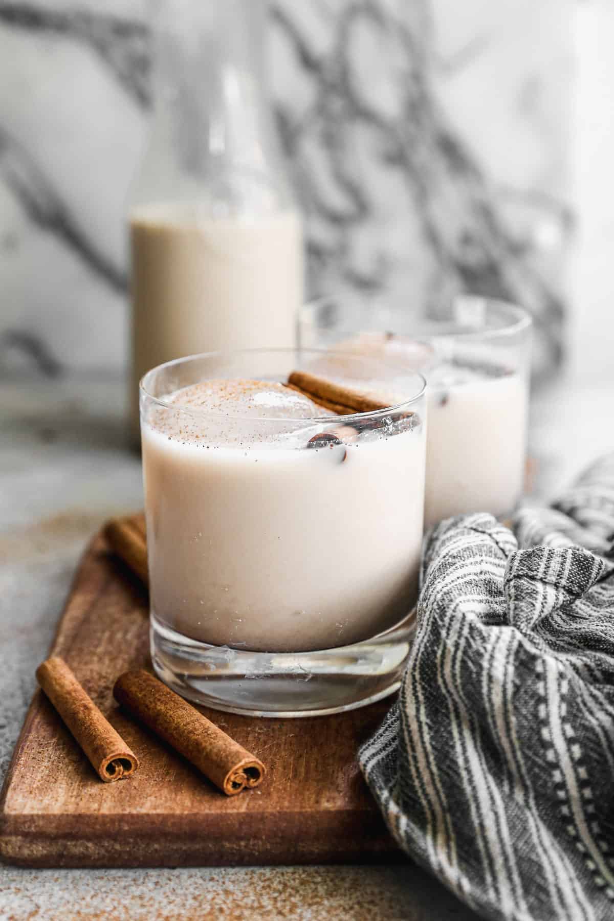 Two glasses of Horchata topped with two cinnamon sticks and a dusting of cinnamon and a bottle behind it.