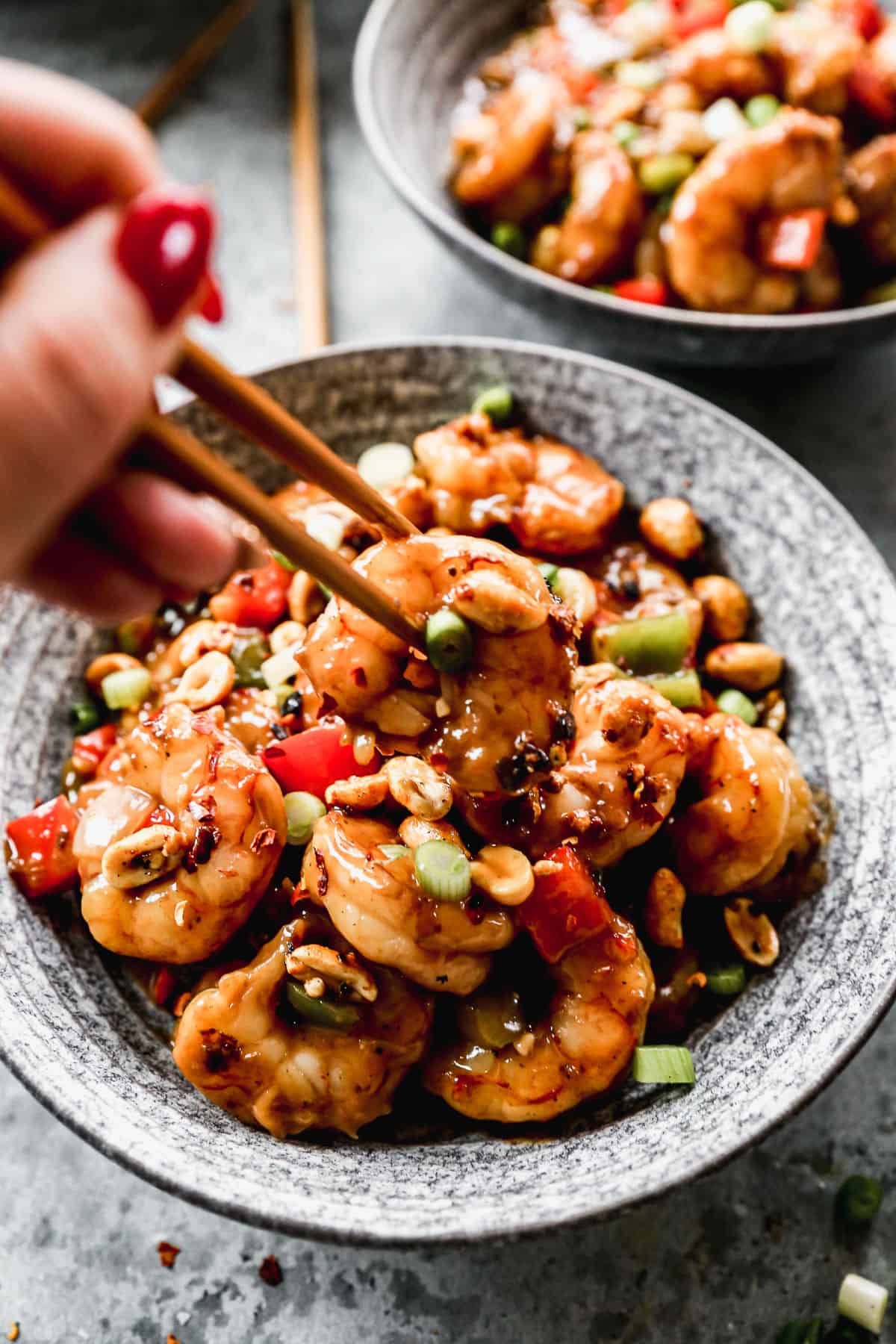 A close-up image of someone lifting a piece of shrimp with chopsticks from a bowl of Kung Pao Shrimp.