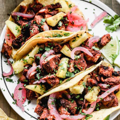 Three Tacos Al Pastor topped with grilled pineapple, pickled red onions, and chopped cilantro on a white plate.