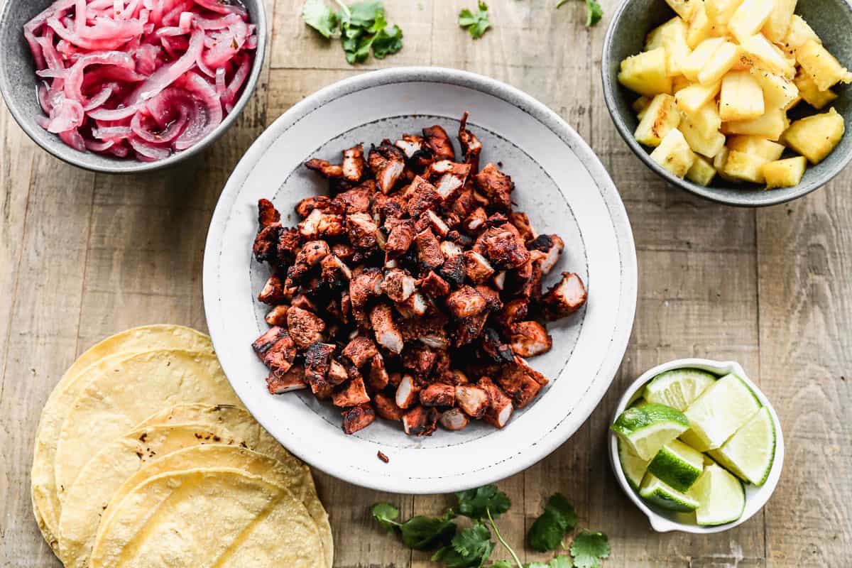 All the foods needed to assemble the best Tacos Al Pastor: corn tortillas, pickled red onion, pork, grilled pineapple, and sliced lime.