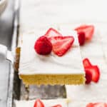 A square of the best White Sheet Cake being lifted from a pan, with a raspberry and sliced strawberries on top.