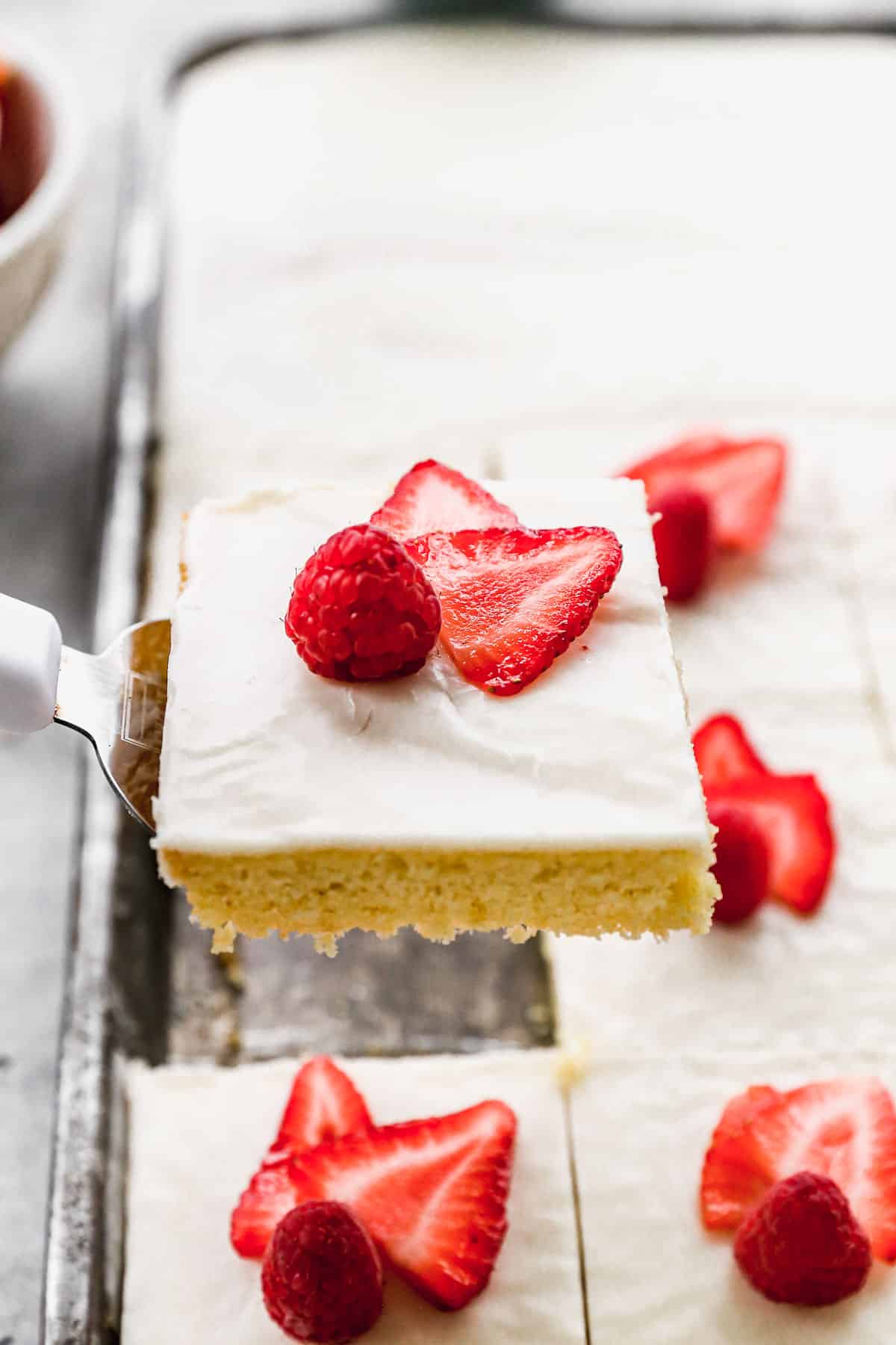 A slice of easy White Texas Sheet Cake being lifted from the pan, topped with a raspberry and sliced strawberries.