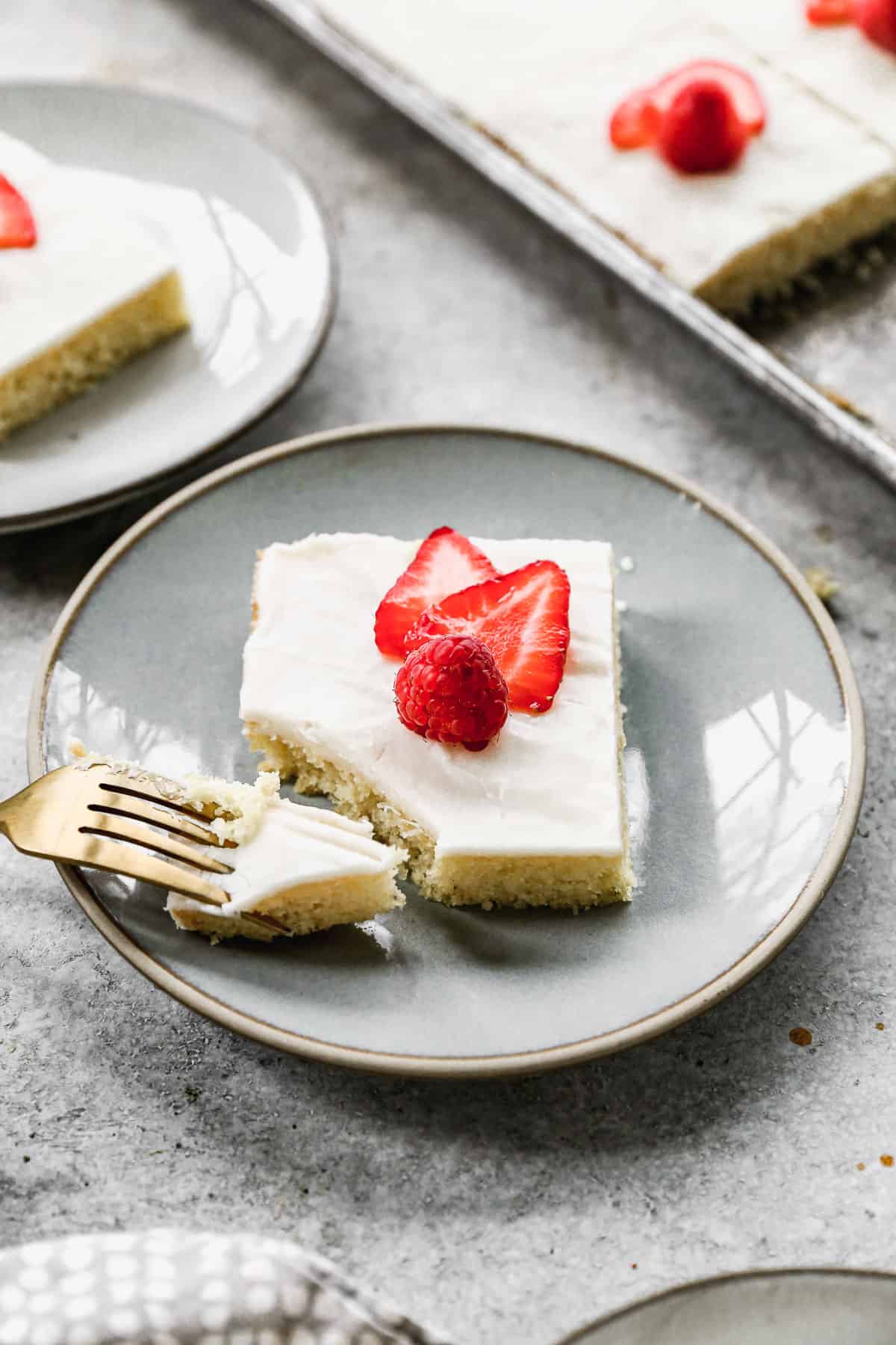 A fork taking a bite from a square of easy White Sheet Cake with white frosting and sliced strawberries and a raspberry on top