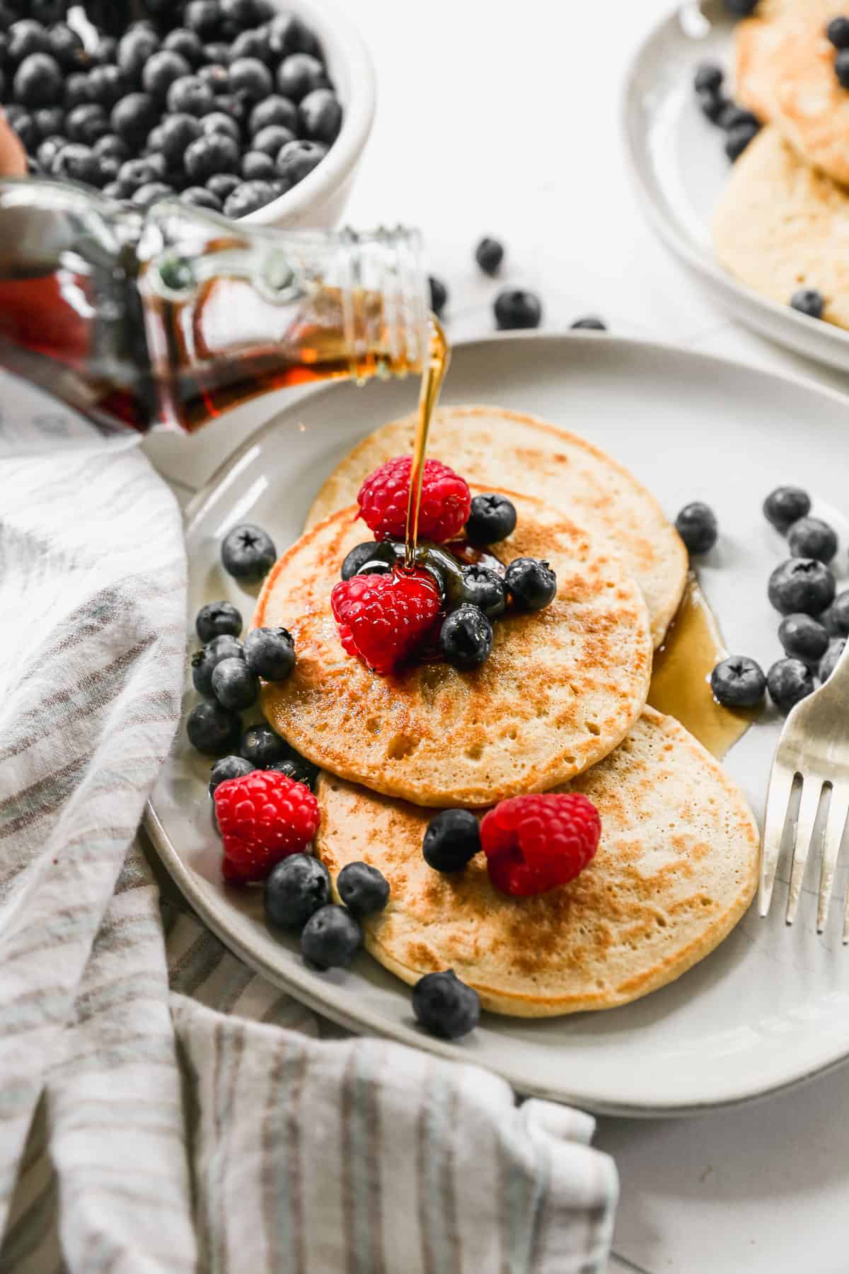 Three Cottage Cheese Pancakes on a plate with blueberries and raspberries, and maple syrup being poured on the top.