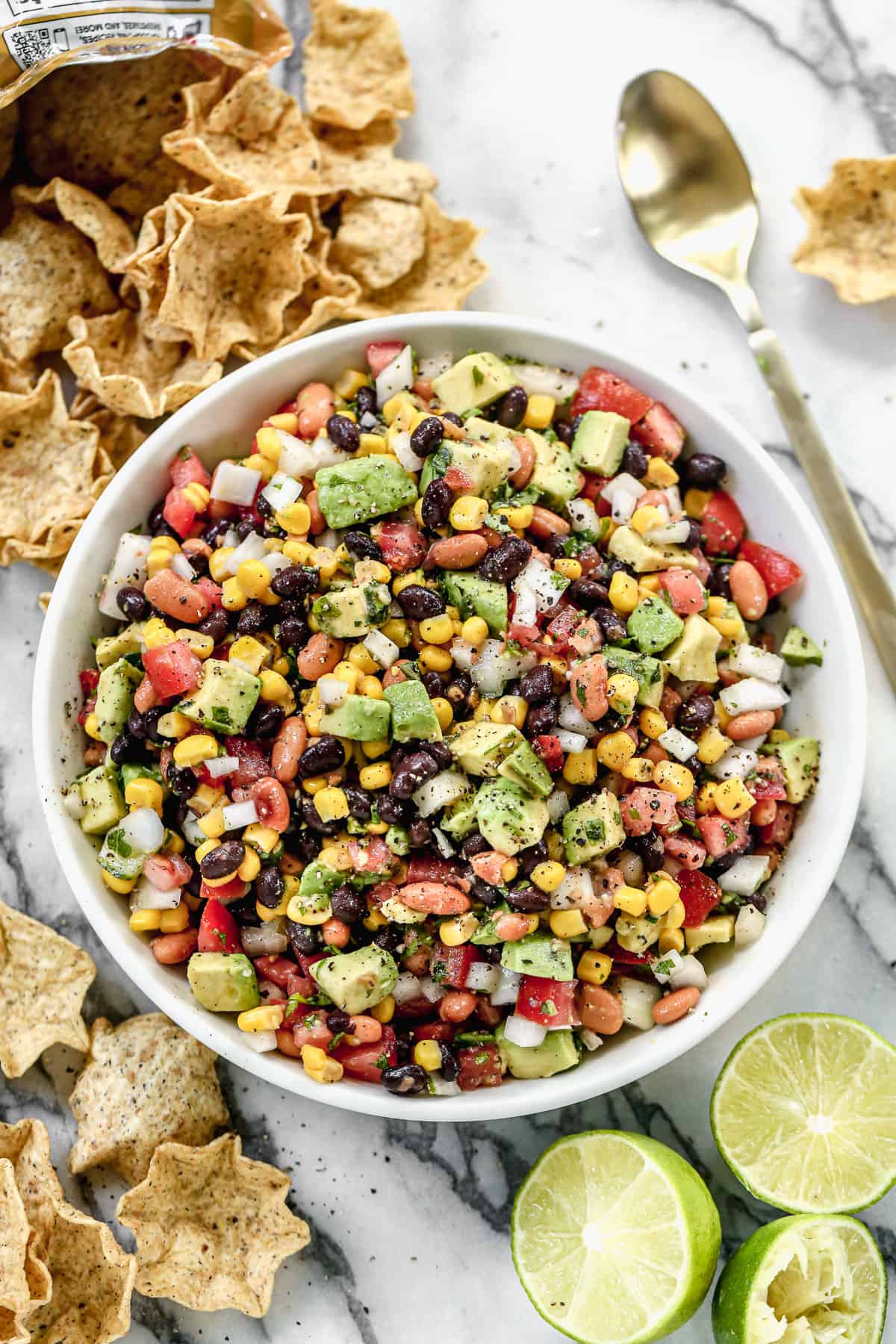 A white serving bowl filled with Cowboy Caviar with chips and lime scattered around it.