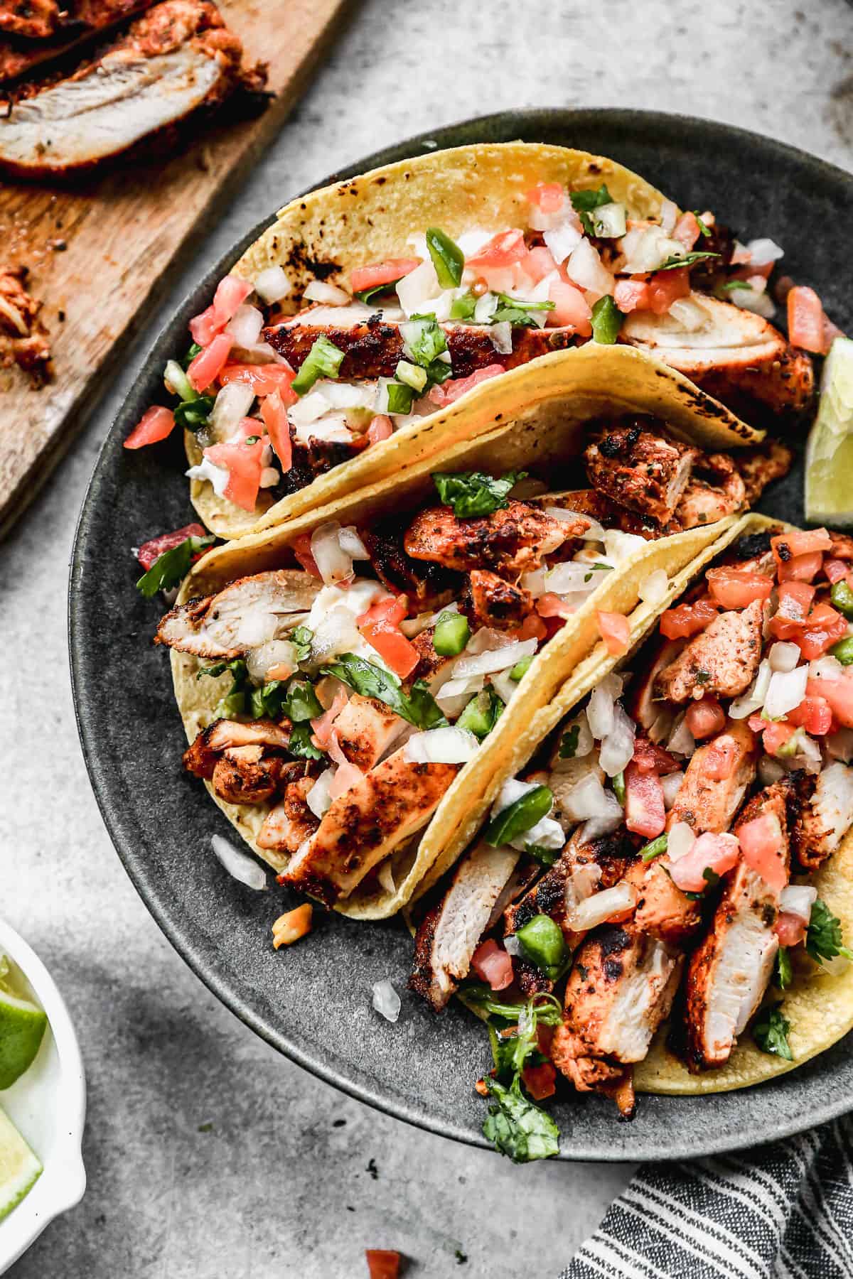 Three grilled chicken street tacos on a plate topped with fresh pico, cilantro, and next to a lime wedge. 