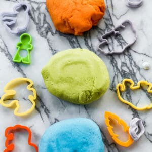 Three balls of homemade playdough on a counter surrounded by playdough cookie cutters. The colors are orange, green, and blue.