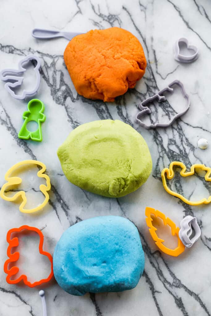 Three balls of homemade playdough on a counter surrounded by playdough cookie cutters. The colors are orange, green, and blue.