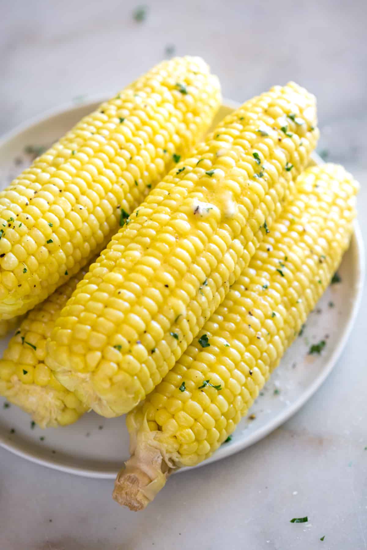 A few corn on the cobs stacked on a yellow plate and drizzled with butter and parsley after being cooked in the Instant Pot.