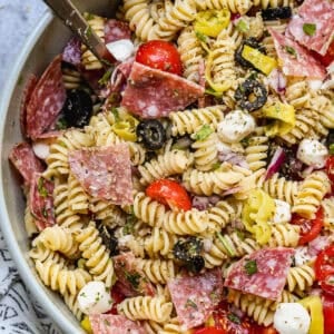 A zesty Italian Pasta Salad recipe served in a large bowl with rotini pasta, mozzarella cheese pearls, salami, black olives, cherry tomatoes, and parmesan cheese.