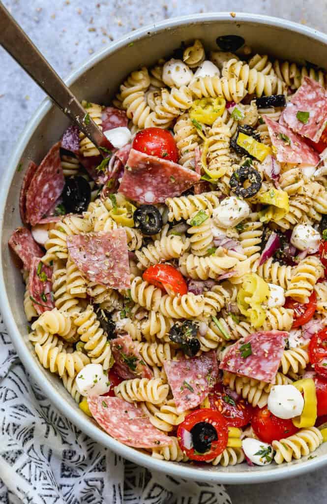 A zesty Italian Pasta Salad recipe served in a large bowl with rotini pasta, mozzarella cheese pearls, salami, black olives, cherry tomatoes, and parmesan cheese.
