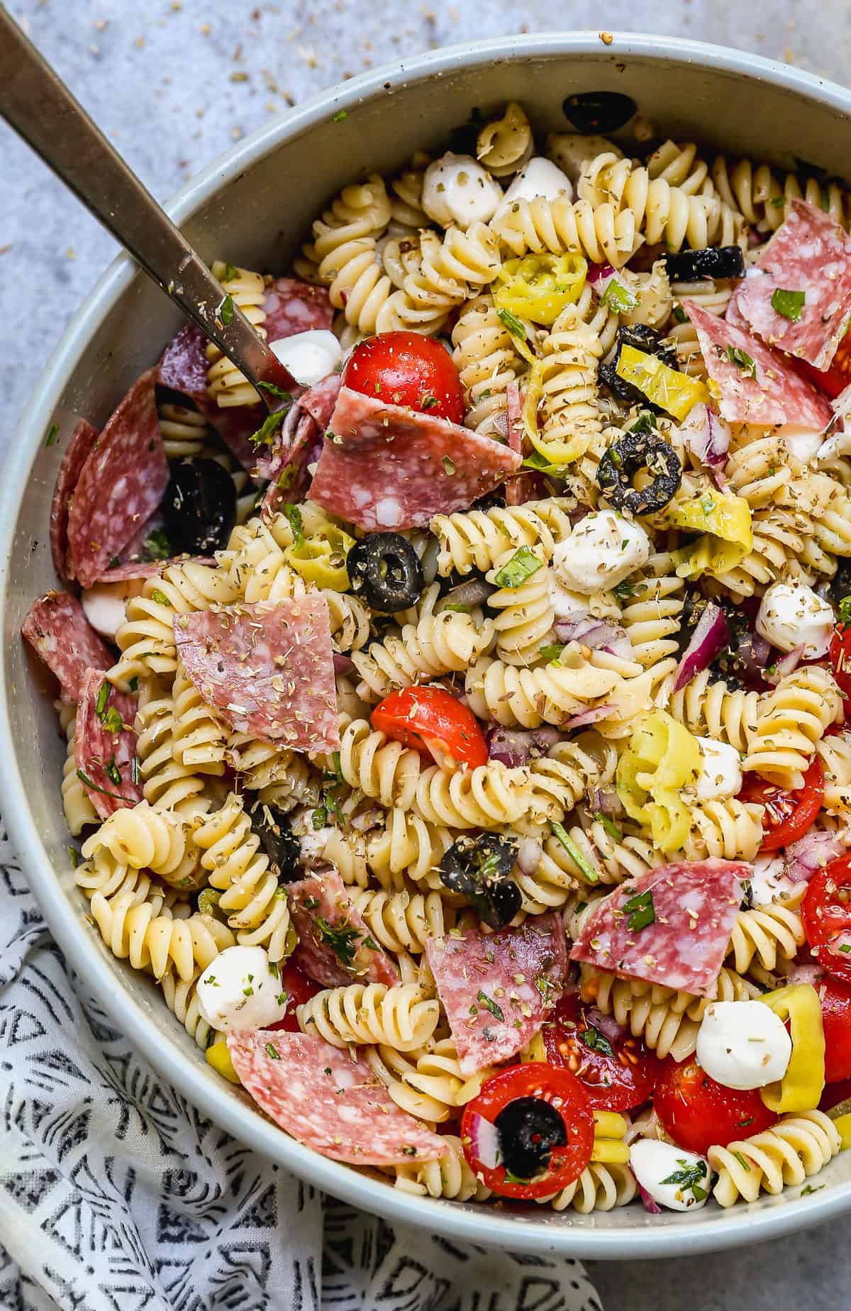 A zesty Italian Pasta Salad recipe served in a large bowl with rotini pasta, mozzarella cheese pearls, salami, black olives, cherry tomatoes, and parmesan cheese.