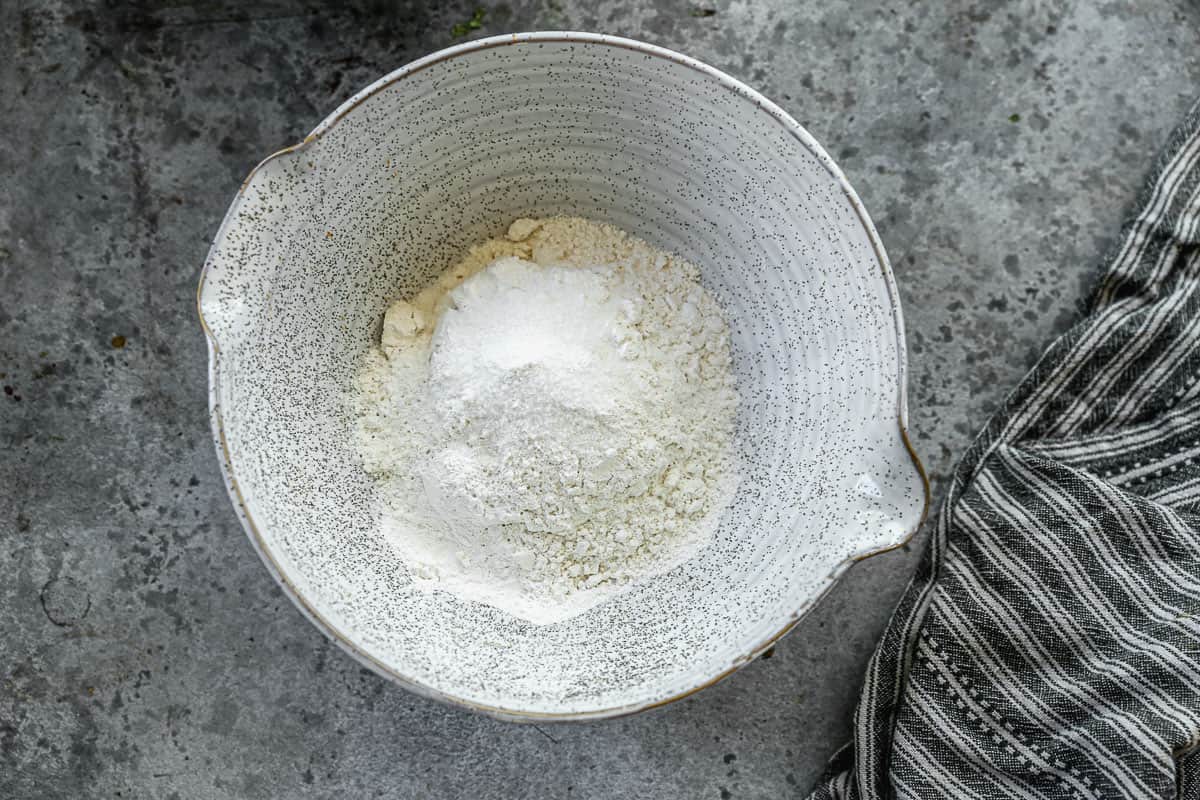 A white stone mixing bowl with flour, baking powder, and salt to make easy Lemon Blueberry Bread.