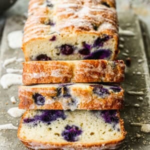 A loaf of the best Lemon Blueberry Bread recipe drizzled with a glaze and half of it sliced.
