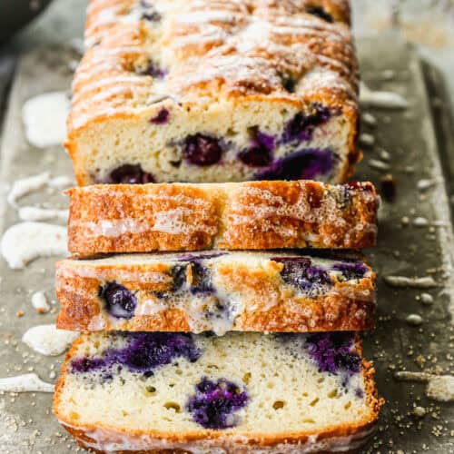 A loaf of the best Lemon Blueberry Bread recipe drizzled with a glaze and half of it sliced.