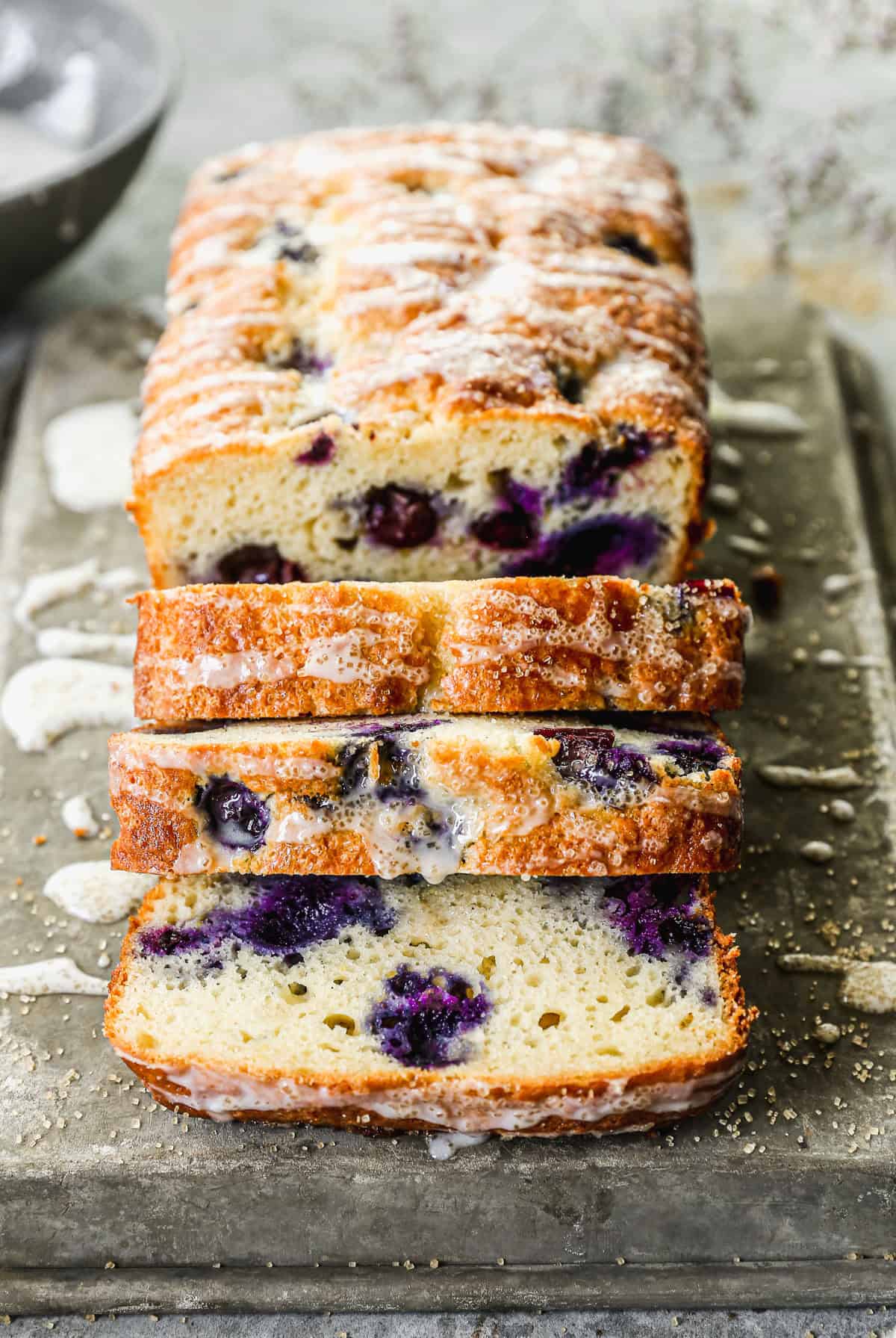 A loaf of the best Lemon Blueberry Bread recipe drizzled with a glaze and half of it sliced.