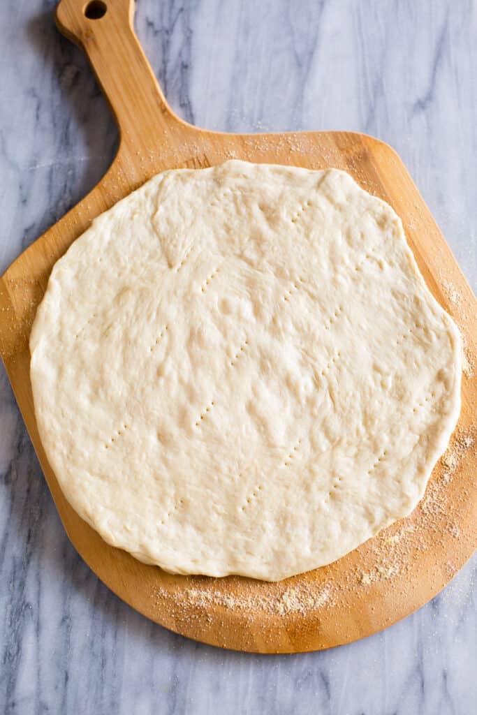 Pizza dough stretched into a round circle and pricked with a fork on a wooden pizza peel.