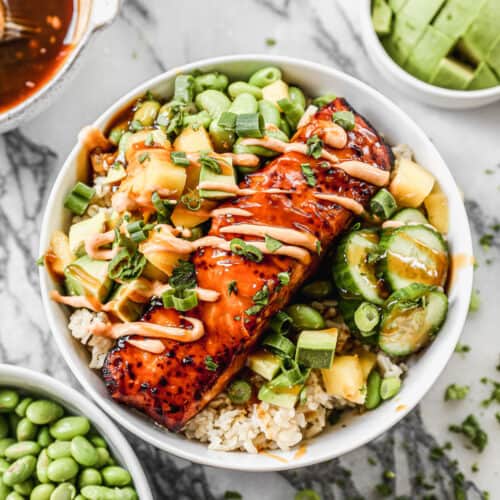 A salmon fillet on a bed of jasmine rice with edamame, mango, avocado, cucumber, green onion, and a drizzle of sriracha mayo.