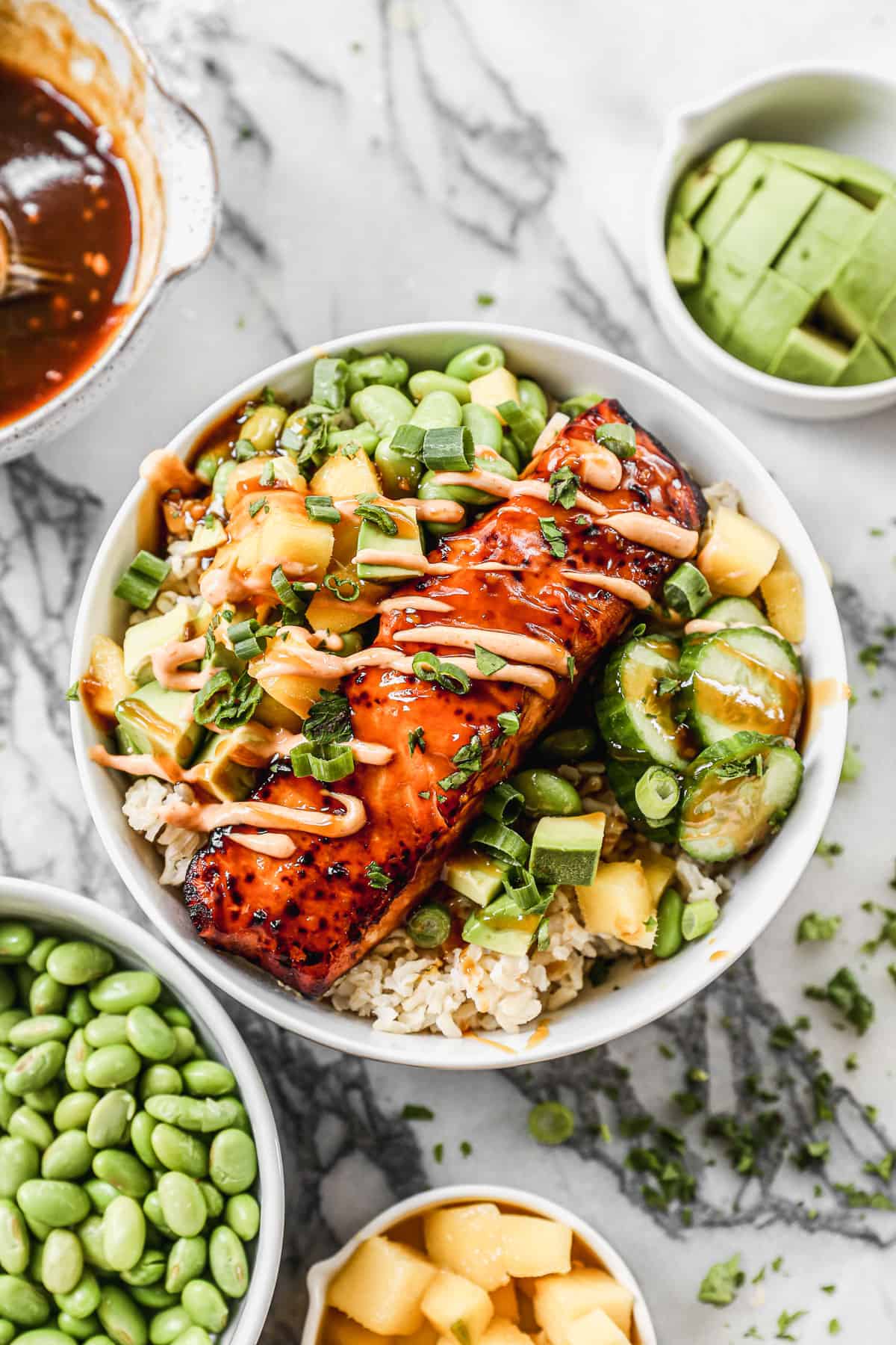 A salmon fillet on a bed of jasmine rice with edamame, mango, avocado, cucumber, green onion, and a drizzle of sriracha mayo.