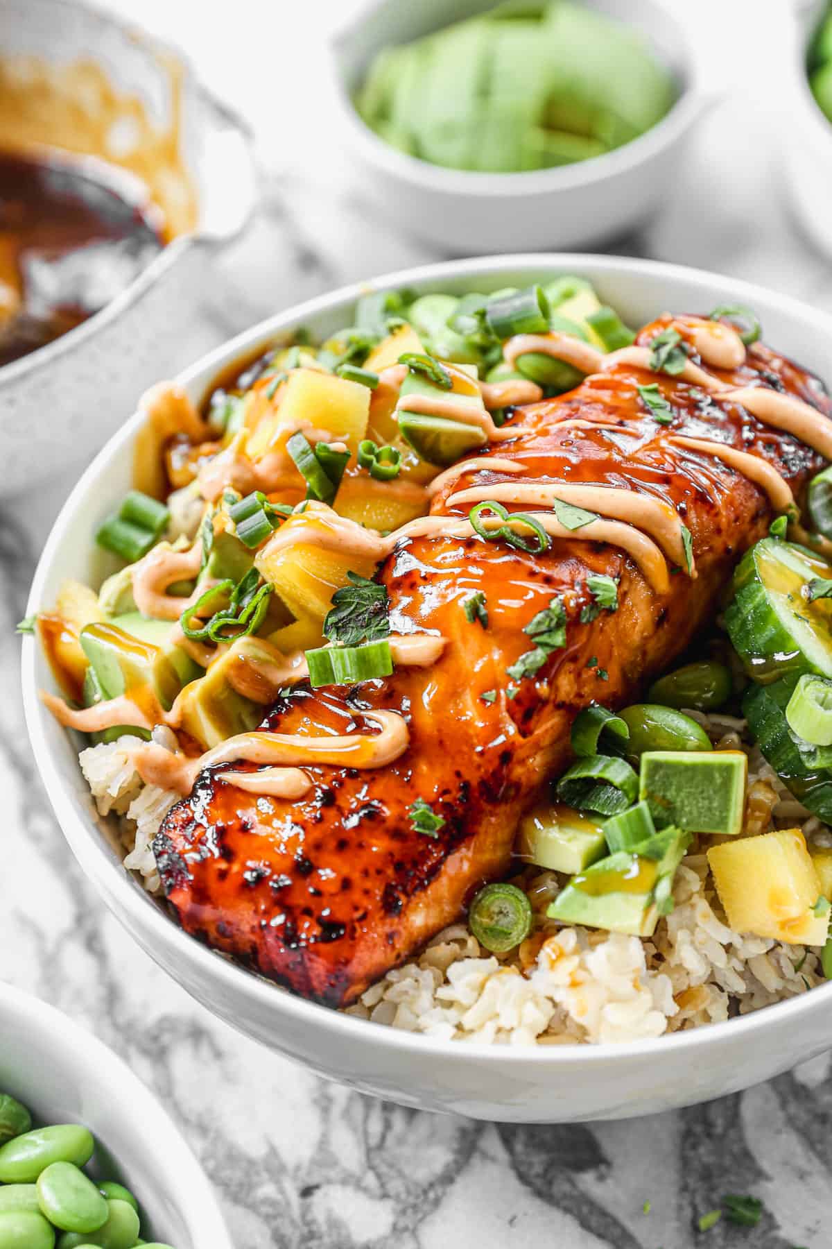 A close-up picture of a salmon bowl with a fillet of teriyaki salmon on top of a bed of jasmine rice with sliced cucumber, mango, green onions, edamame, and avocado. 