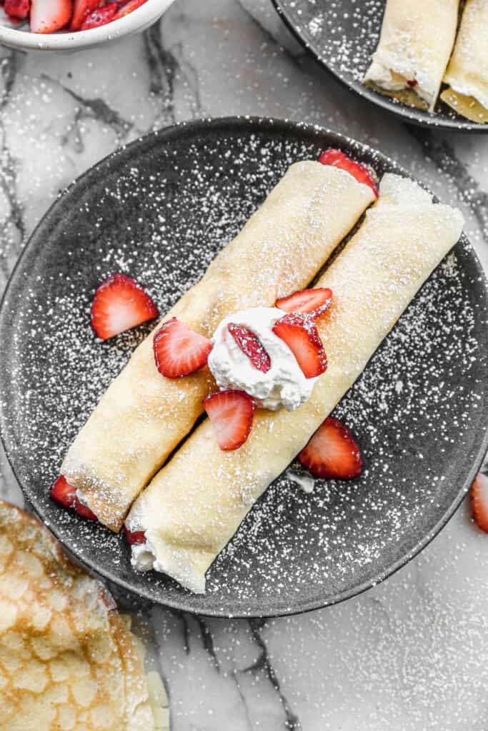 Two homemade Strawberry Crepes topped with whipped cream, strawberries, and a dusting of powdered sugar.