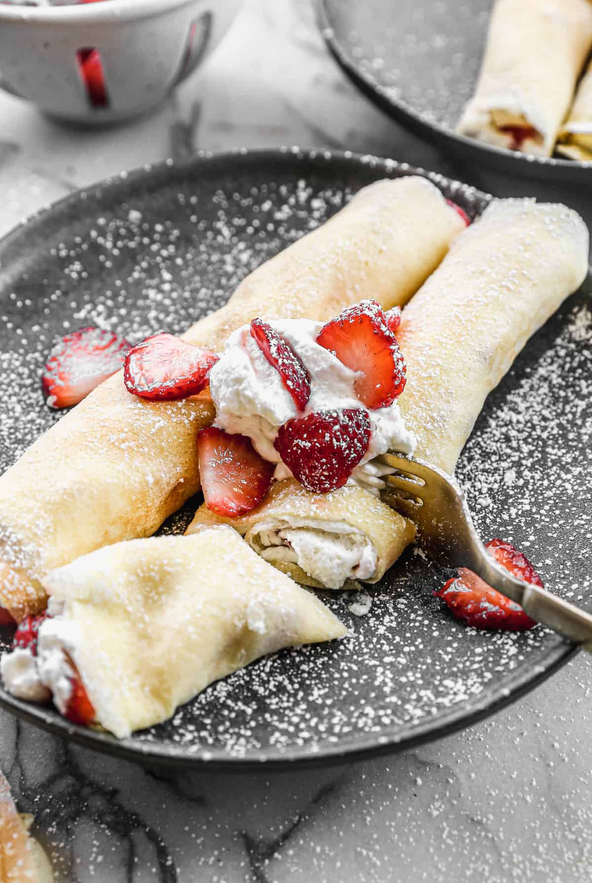 A close up image of a fork cutting into an easy Strawberry Crepe topped with whipped cream, powdered sugar, and strawberries.