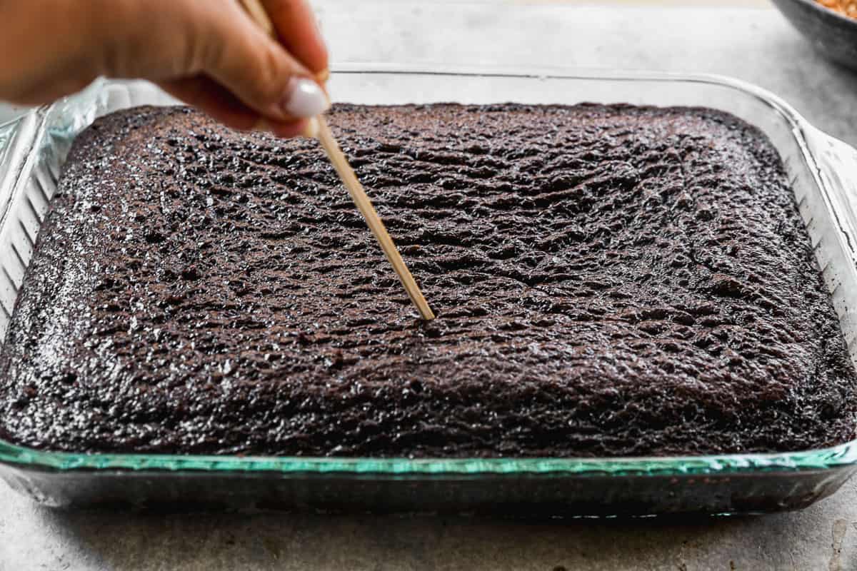 A chocolate cake in a 9x13 pan being poked with a wooden skewer. 