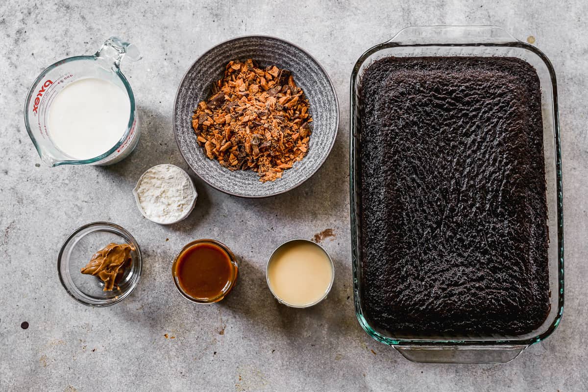 All the ingredients needed to make the best Butterfinger Cake: chocolate cake, crushed butterfingers, sweetened condensed milk, caramel sauce, peanut butter, heavy cream, and powdered sugar.