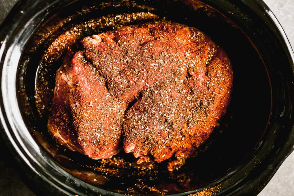 A pork shoulder roast in a slow cooker with seasoning on all side to make easy carnitas.