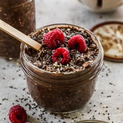 A Chocolate Peanut Butter Overnight Oats recipe in a half pint jar with a wooden spoon, topped with cacao nibs and fresh rasberries.