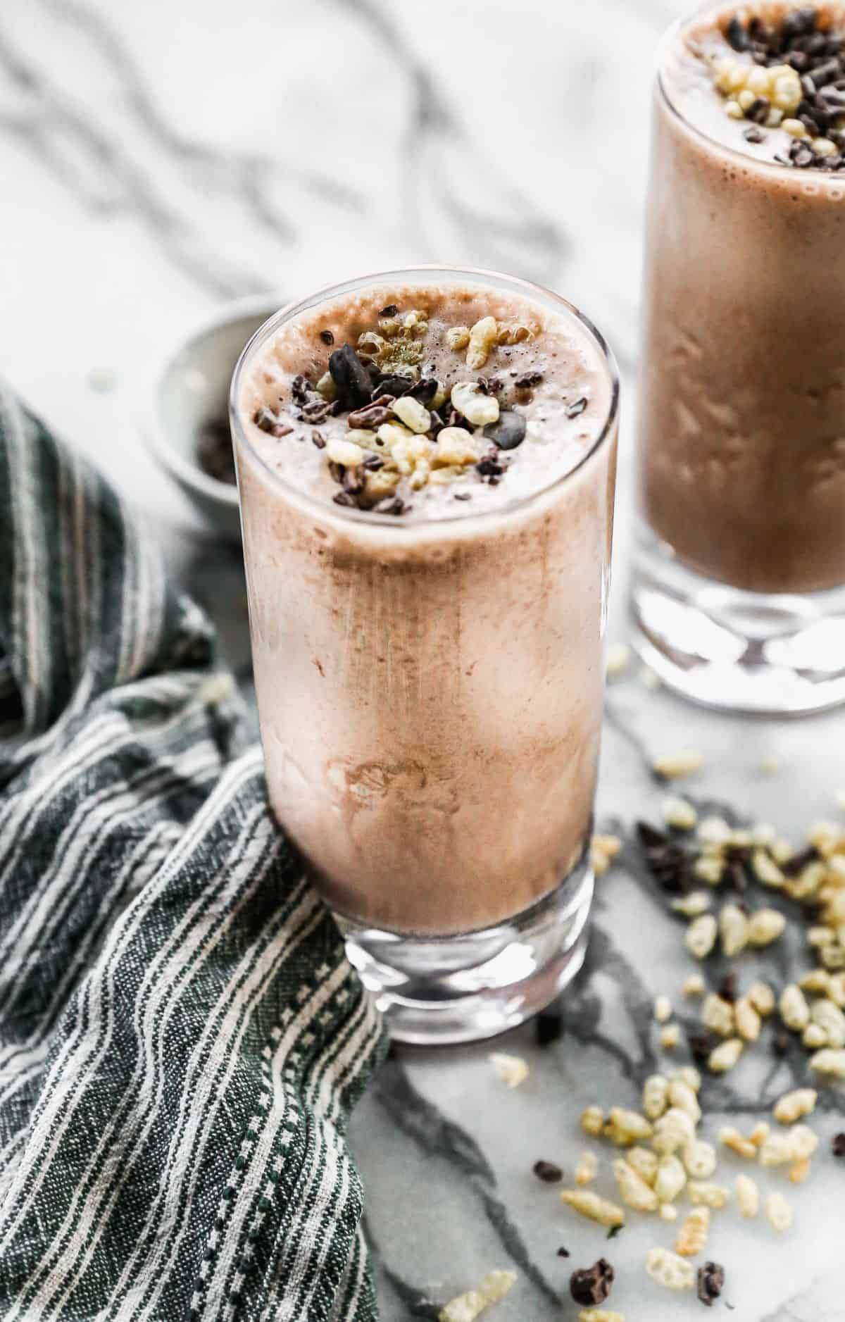 A chocolate protein shake in a tall glass and topped with cocoa nibs and rice crispy cereal.