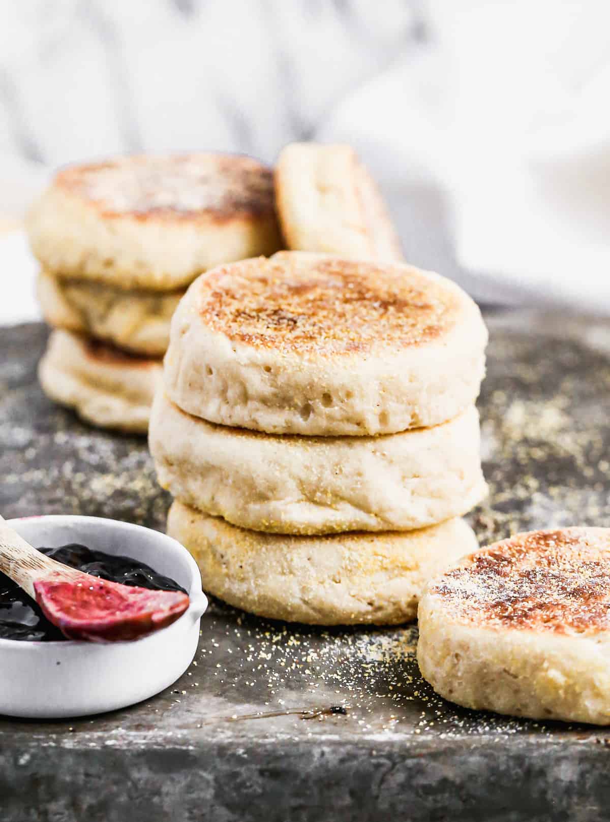 Three homemade English Muffins stacked on each other on a baking sheet next to a cup of jam.