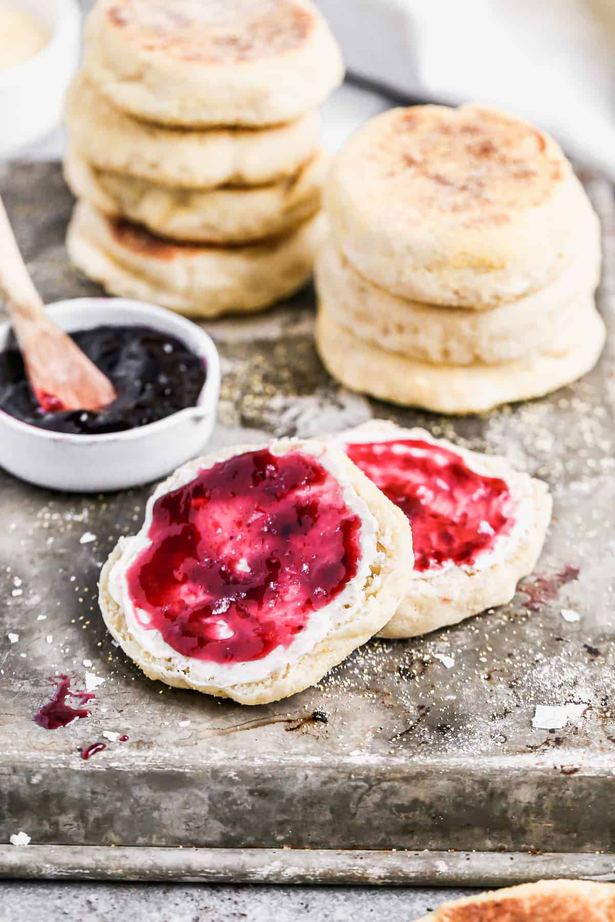 A homemade English Muffin split in half and spread with butter and jam, with a stack of more english muffins behind.
