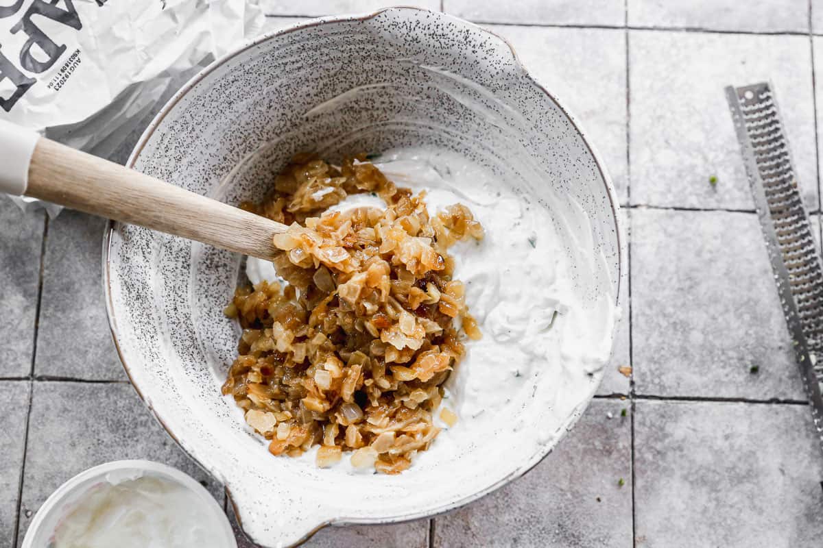 A mixing bowl with caramelized onions being stirred into a greek yogurt base for an easy French Onion Dip.
