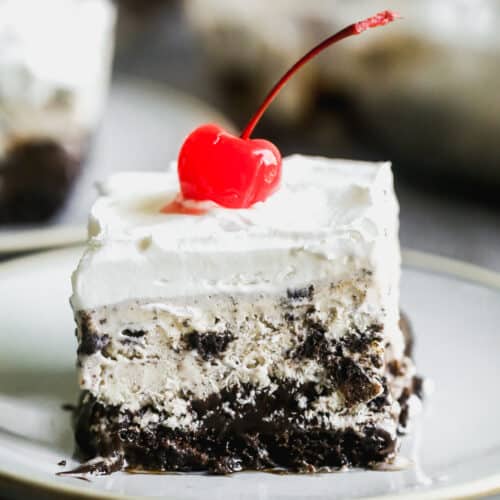 A piece of an easy Ice Cream Cake made with chocolate cake, cookies and cream ice cream, hot fudge, and topped with whipped cream and a cherry.