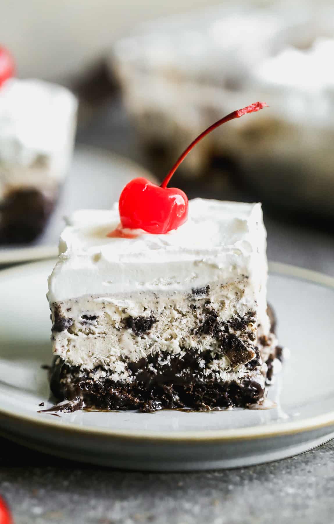 A piece of homemade ice cream cake with chocolate cake as the base, hot fudge, cookies and cream ice cream, whipped cream, and a cherry on top.
