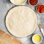 A homemade Pizza Dough recipe rolled out and surrounded by the supplies for a homemade pepperoni pizza.