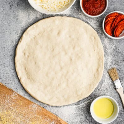 A homemade Pizza Dough recipe rolled out and surrounded by the supplies for a homemade pepperoni pizza.