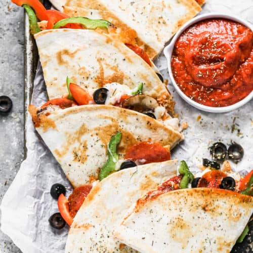 A pizza quesadilla filled with olives and bell peppers on a baking sheet with a cup of marinara sauce for dipping.