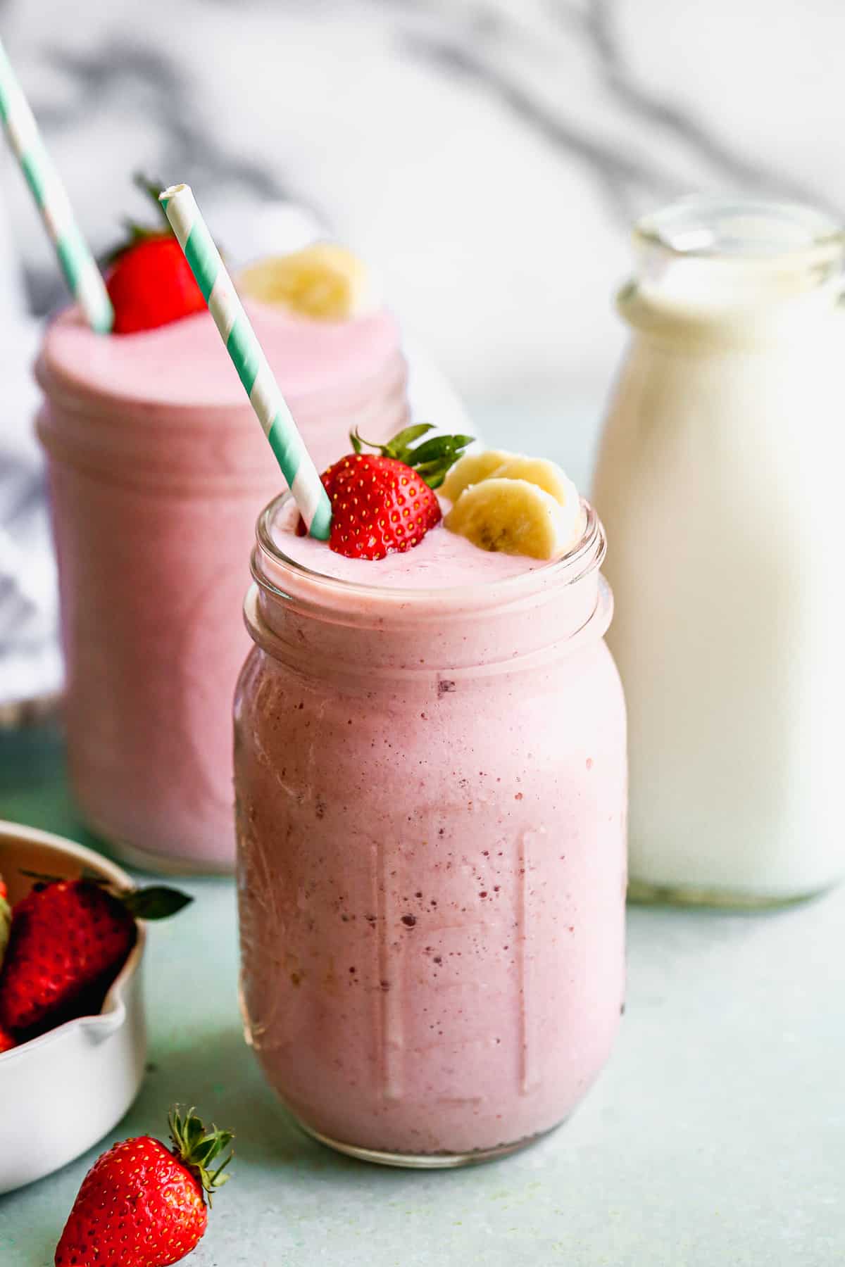 A strawberry banana smoothie in a large mason jar topped with a fresh strawberry and banana slices and a striped straw.