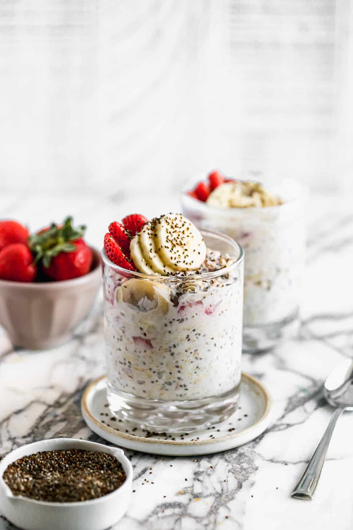 A clear glass filled with Strawberry Overnight Oats and topped with fresh sliced strawberries, bananas, and chia seeds.