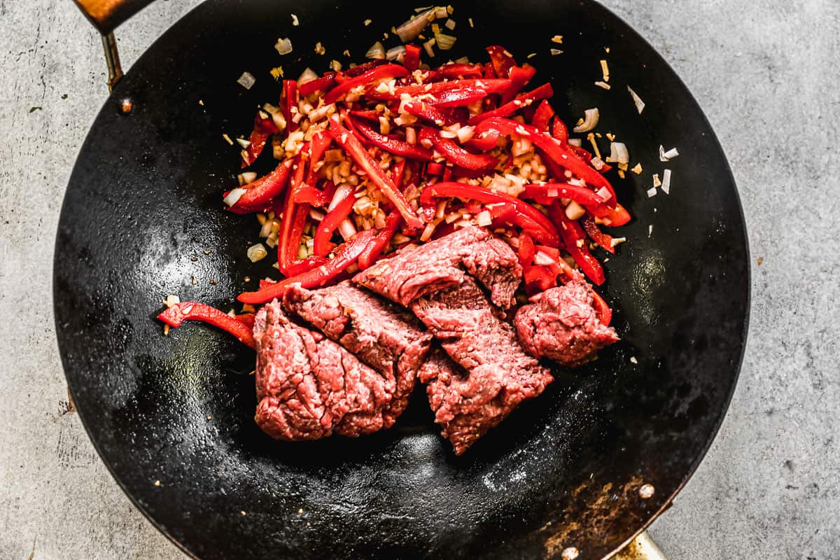 A wok with oil, shallots, garlic, ginger and bell peppers pushed to the side and raw ground beef placed in the pan.