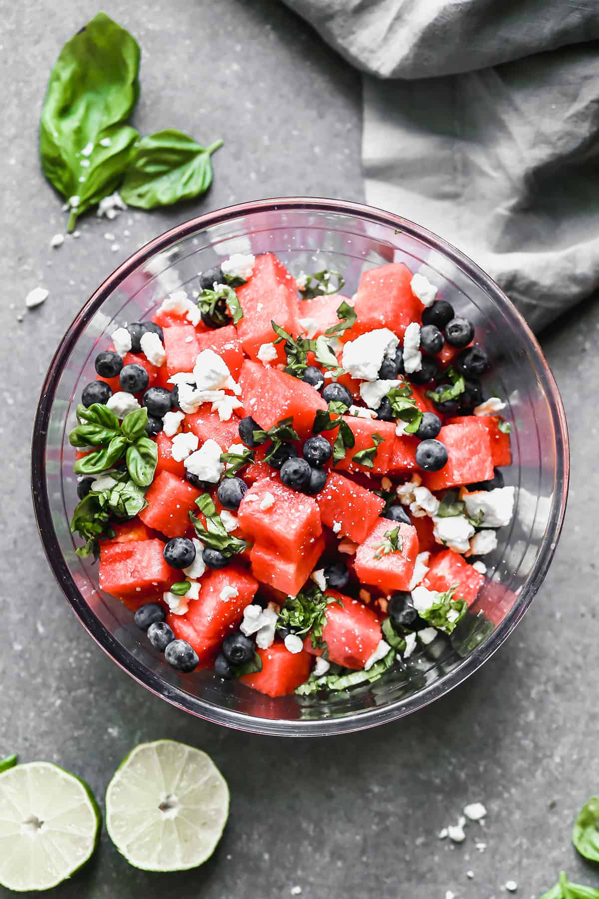 A glass serving bowl filled with fresh watermelon, blueberries, basil, and feta cheese. 