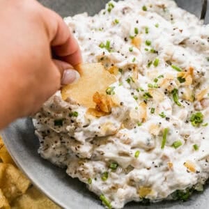 A homemade french onion dip recipe with fresh caramelized onions and herbs, with someone dipping a chip in it.